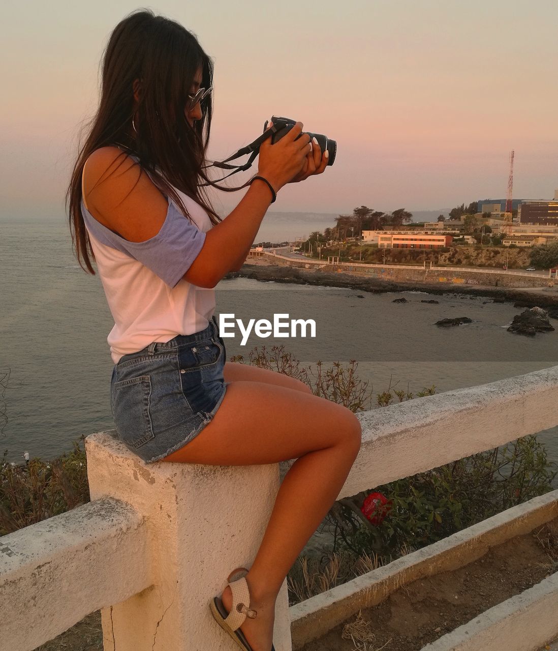 Side view of woman photographing while sitting on railing against sky