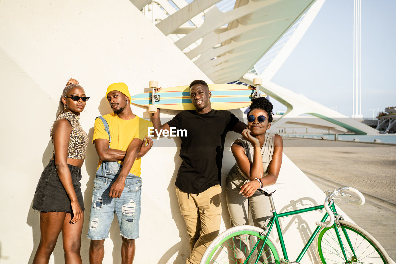 Group of african american friendly people in stylish summer clothes standing together on street with longboard and modern bike while looking at camera