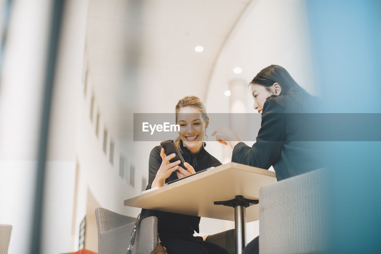 Low angle view of smiling businesswoman sharing smart phone with female colleague at table in office