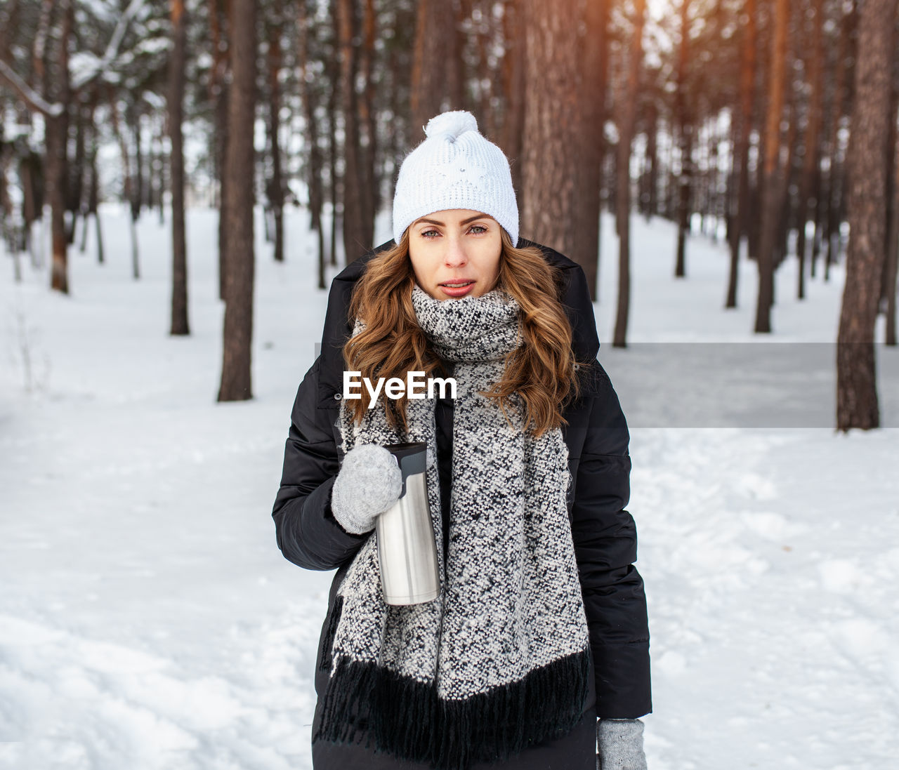 Portrait of confident woman holding insulated drink container while standing in forest during winter