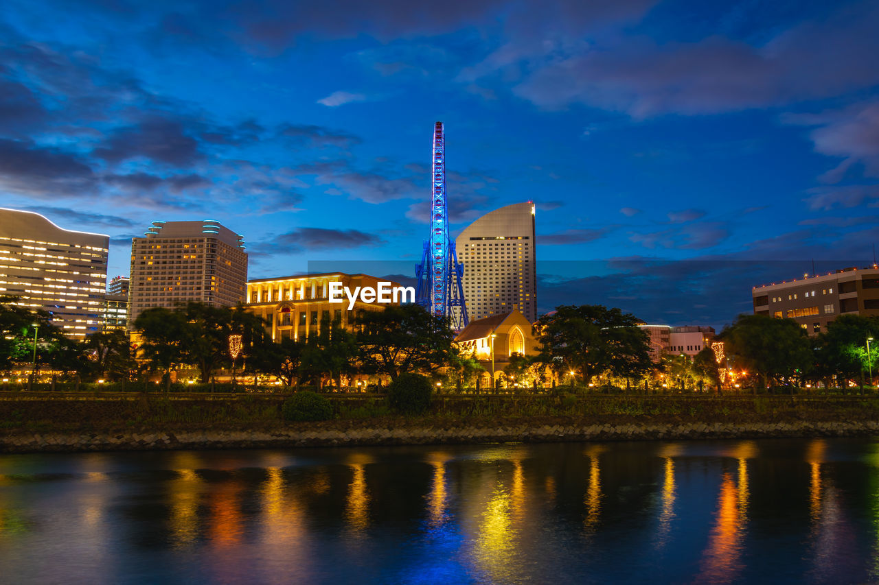 ILLUMINATED BUILDINGS BY RIVER AGAINST SKY AT DUSK