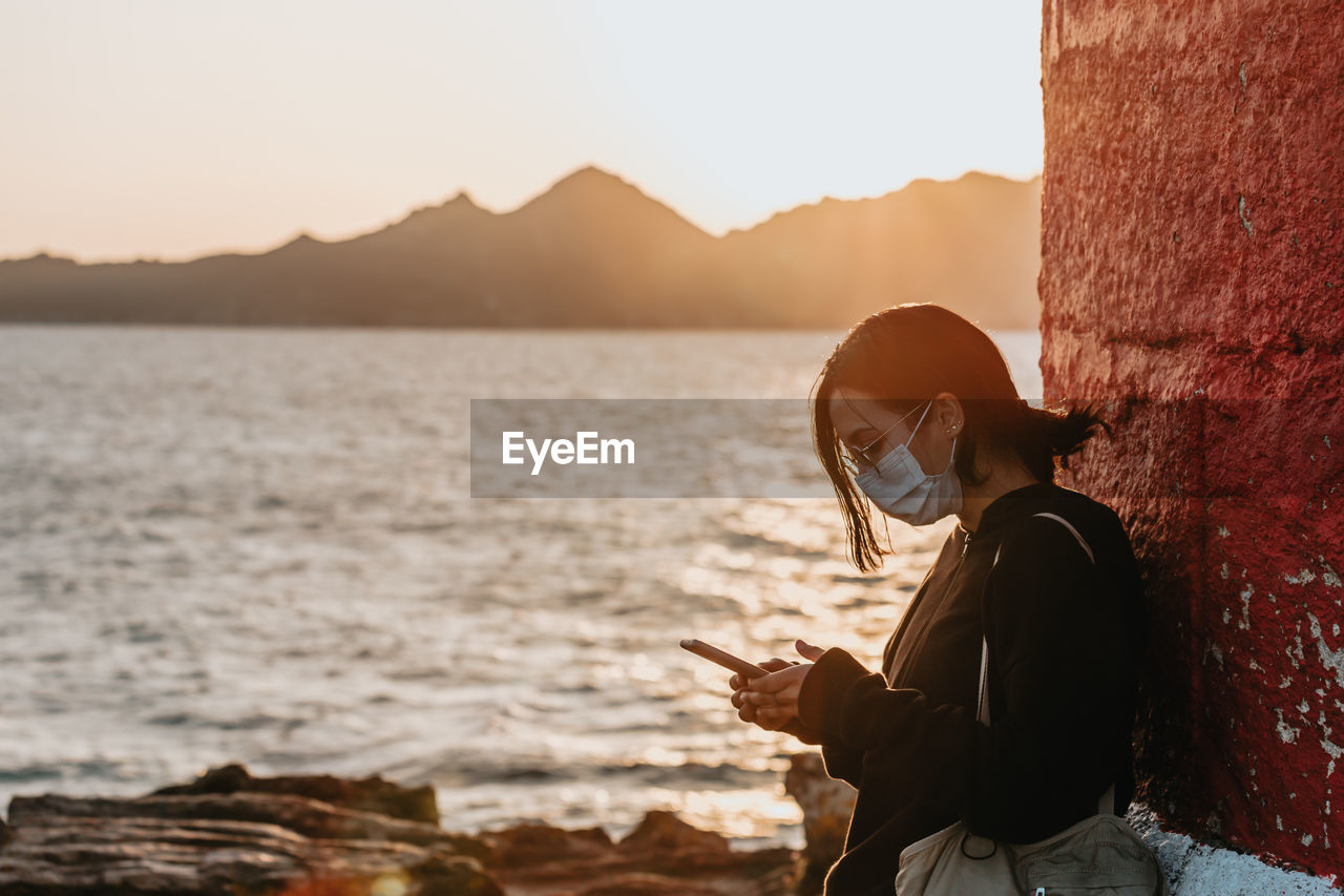 Side view of woman wearing mask using phone against sea