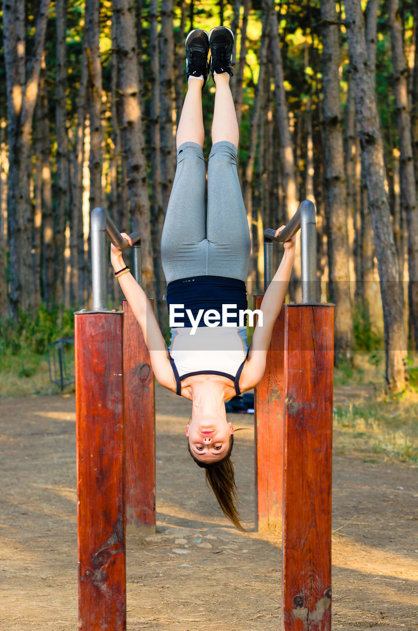Full length of young woman hanging upside down on parallel bars against trees in forest