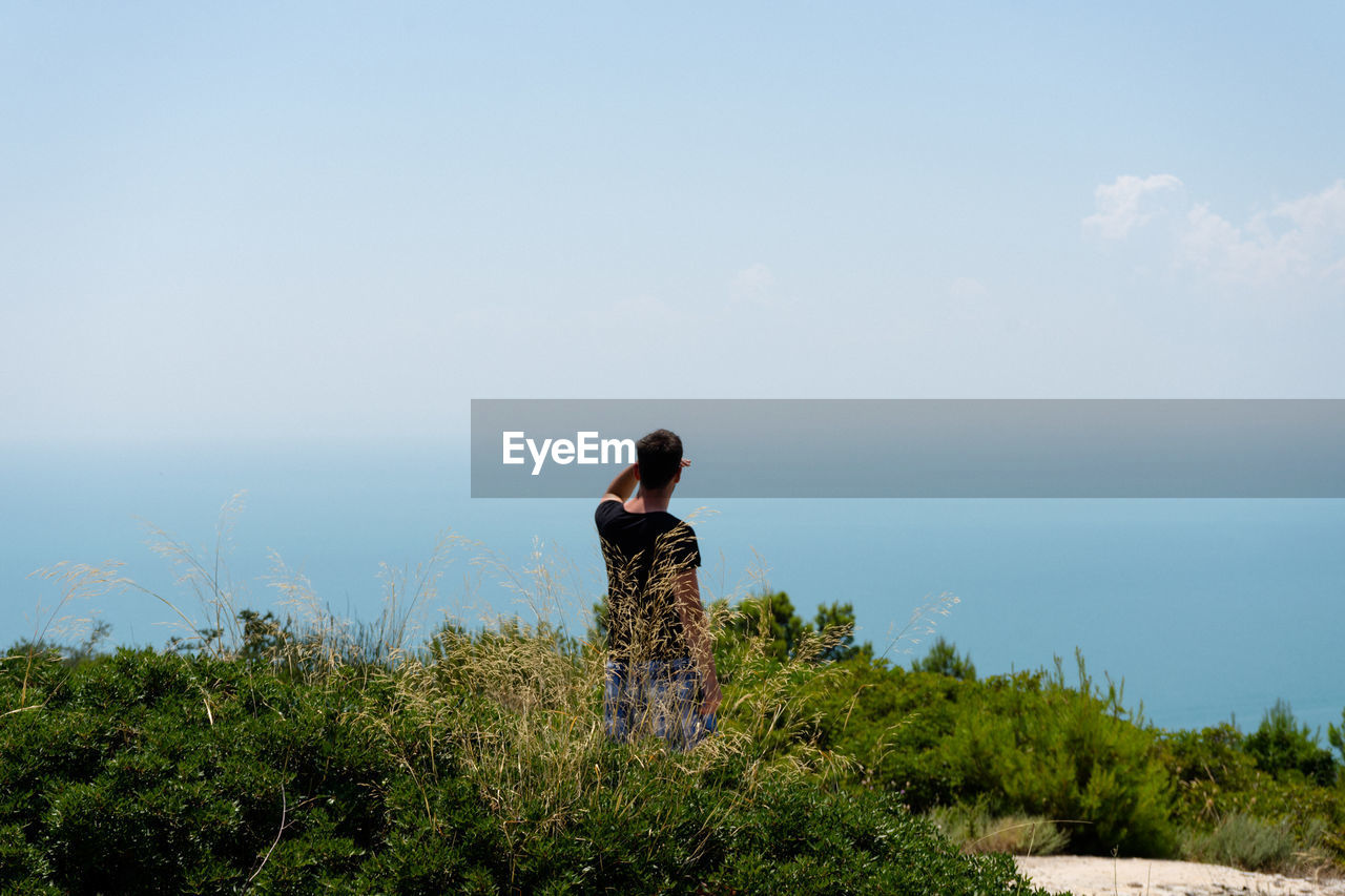 Rear view of man looking at sea while standing amidst plants against sky