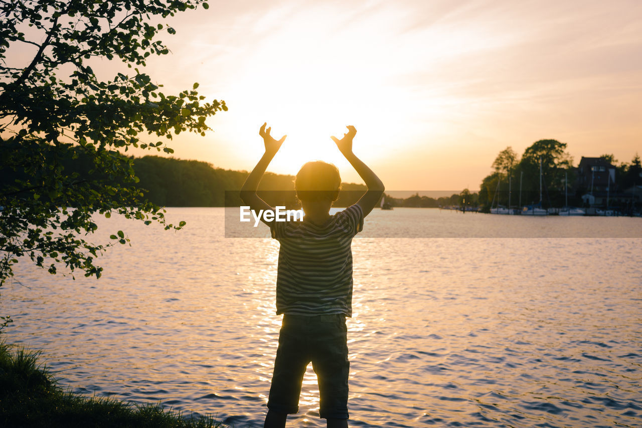 Rear view of boy standing by lake against sky during sunset