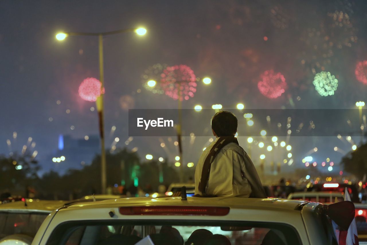 Rear view of boy looking at firework display while sitting on car roof at night