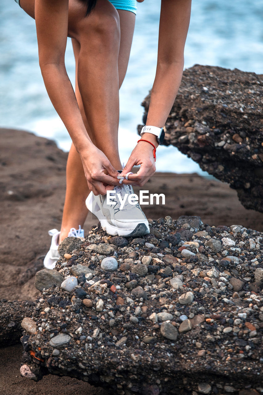 Ethnic female athlete tying shoelaces on sneakers while sitting on seashore and preparing for workout