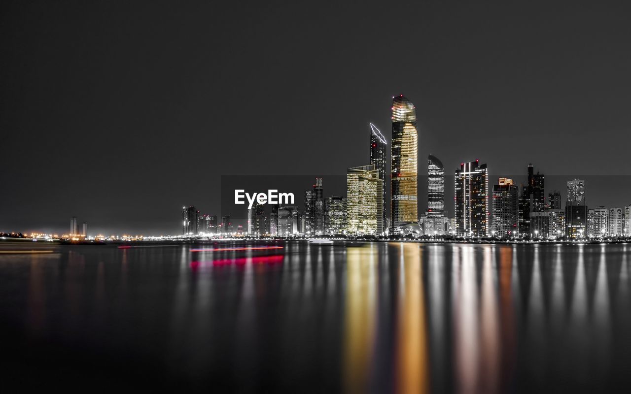 Illuminated buildings by river against sky at night