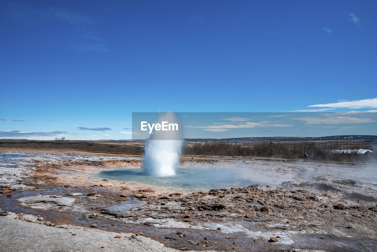 Eruption of strokkur geyser amidst landscape against blue sky during sunny day