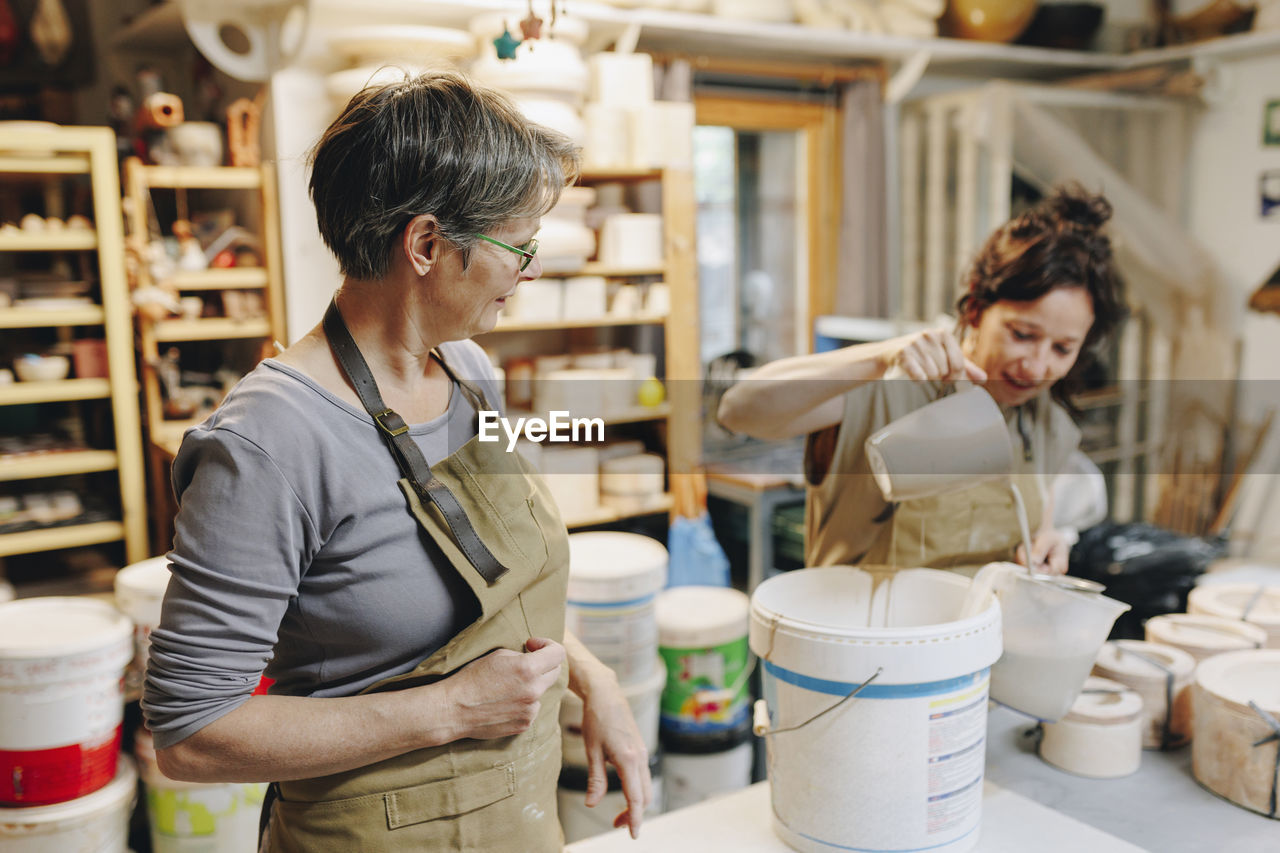 Potter with colleague pouring clay in plastic mug at ceramics workshop