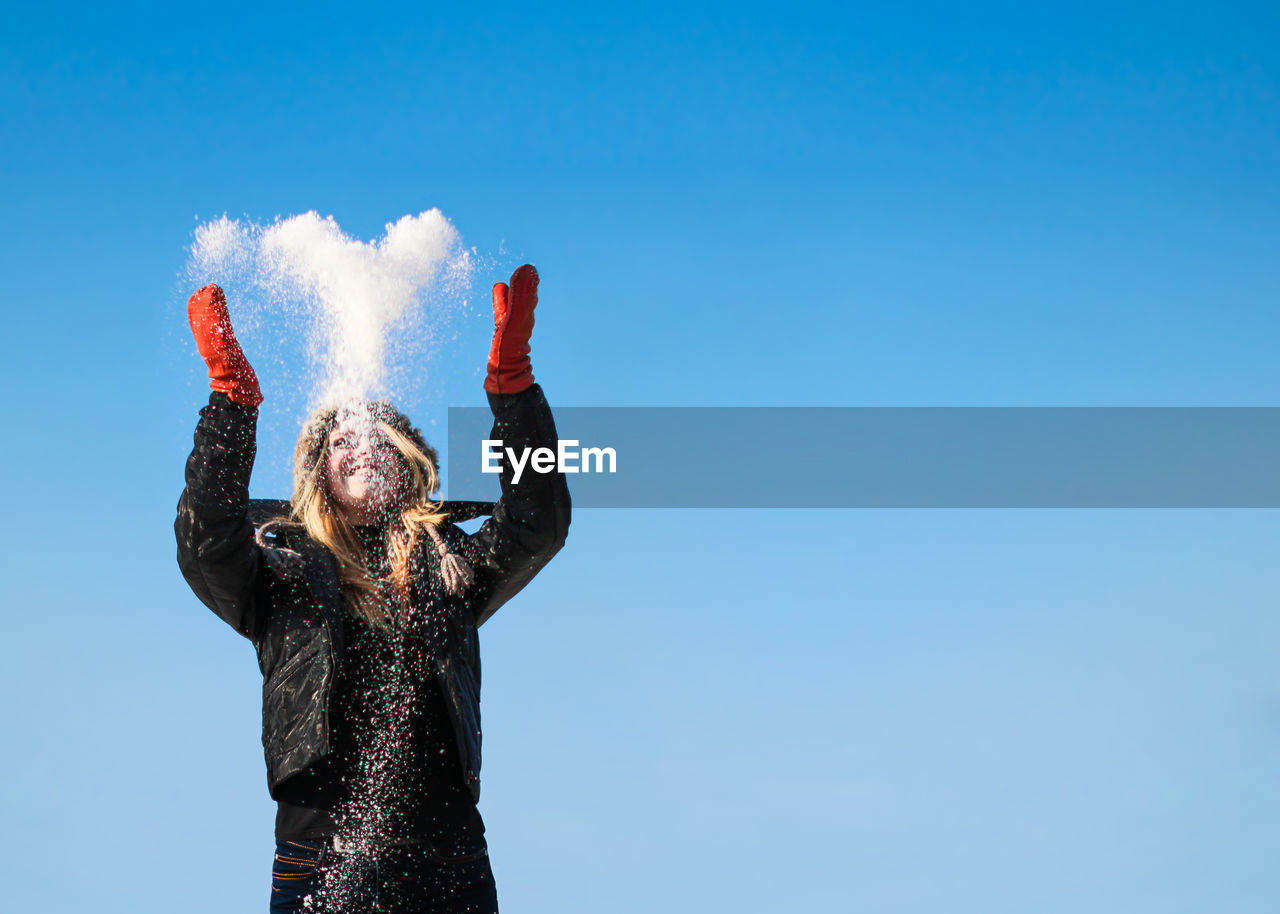 Happy young woman throwing snow against clear blue sky