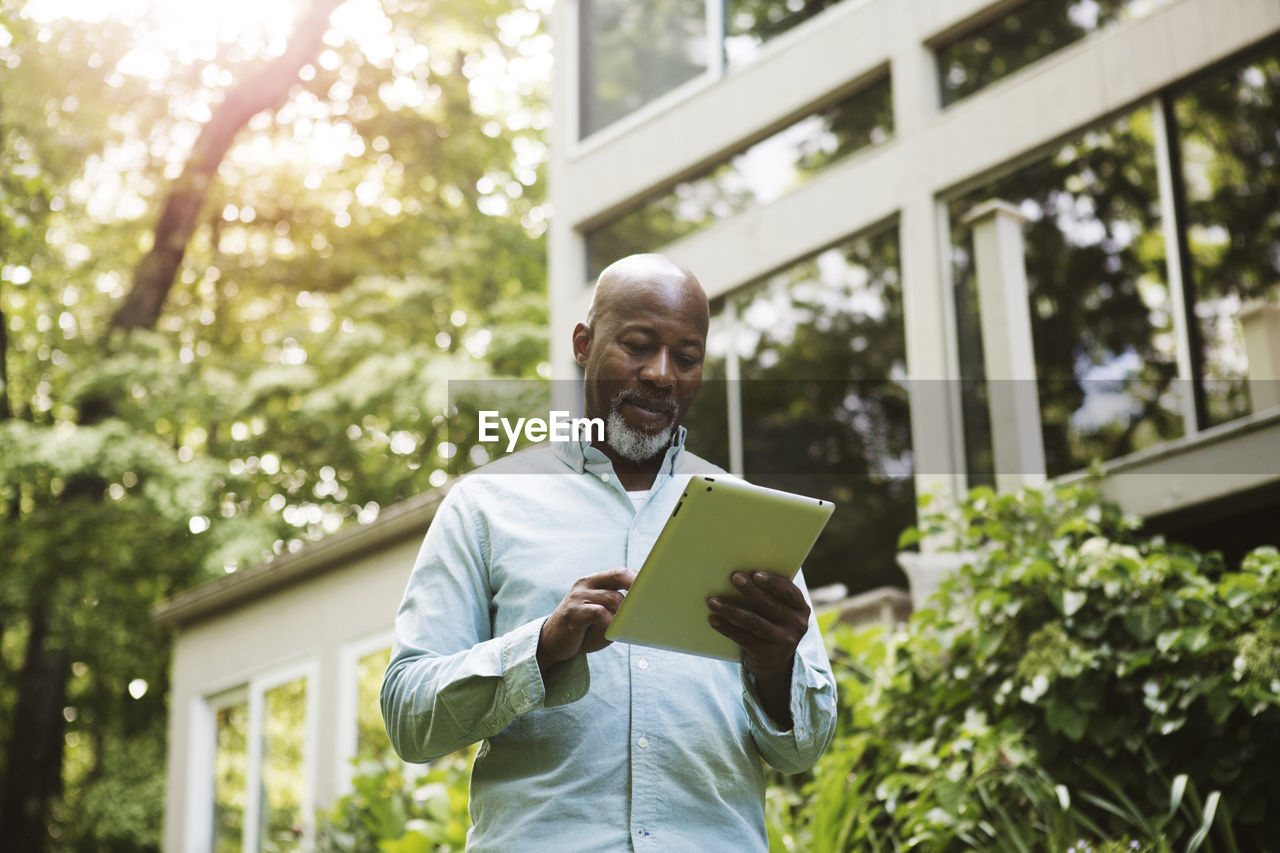 Senior man looking at tablet outside house on sunny day