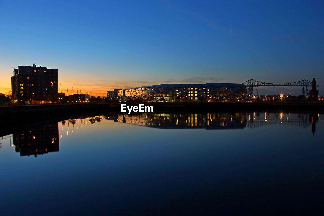 Illuminated buildings by river against sky at dusk