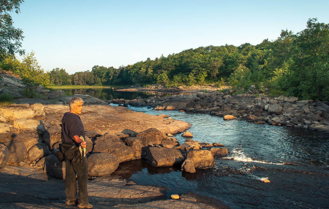 REAR VIEW OF MAN STANDING ON ROCK BY RIVER AGAINST SKY