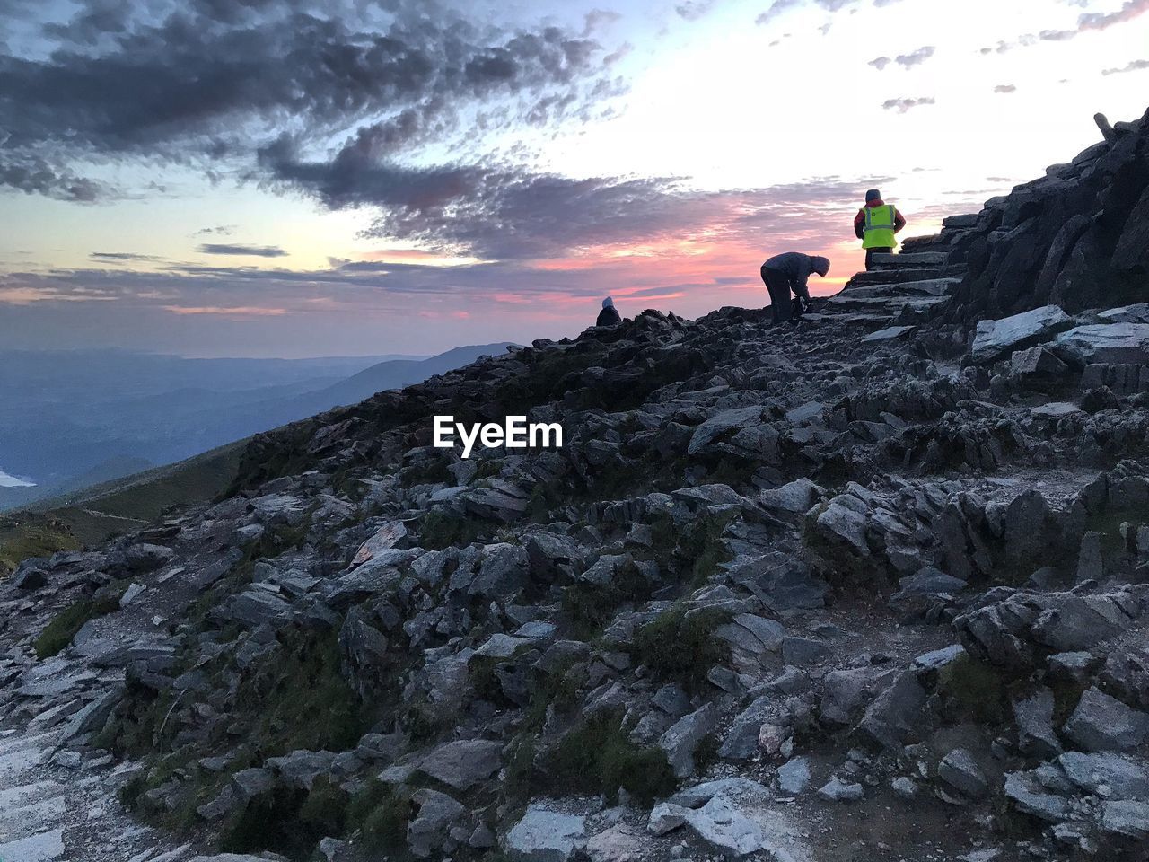 PEOPLE ON ROCKS BY MOUNTAINS AGAINST SKY