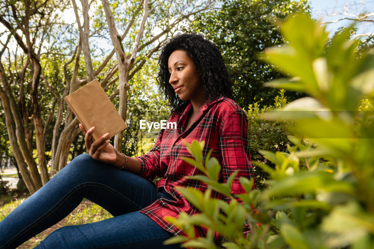 BEAUTIFUL YOUNG WOMAN SITTING ON PLANT AGAINST TREES