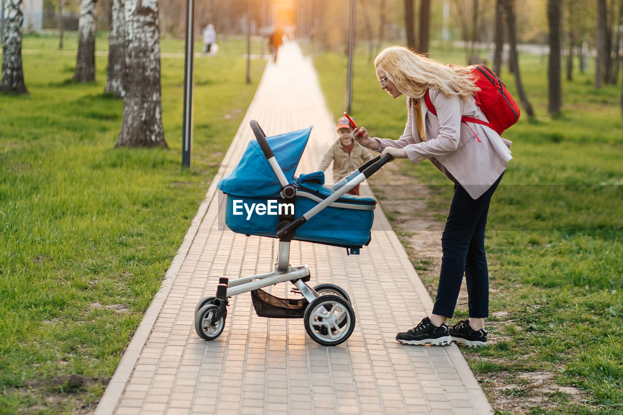 baby carriage, full length, women, adult, one person, transportation, plant, lifestyles, vehicle, nature, grass, casual clothing, lawn, female, park, park - man made space, footpath, leisure activity, tree, day, wheel, cart, outdoors, person, blond hair, clothing, childhood, pushing, young adult, child, activity, side view, mode of transportation, emotion, happiness, bag, sunlight, shopping