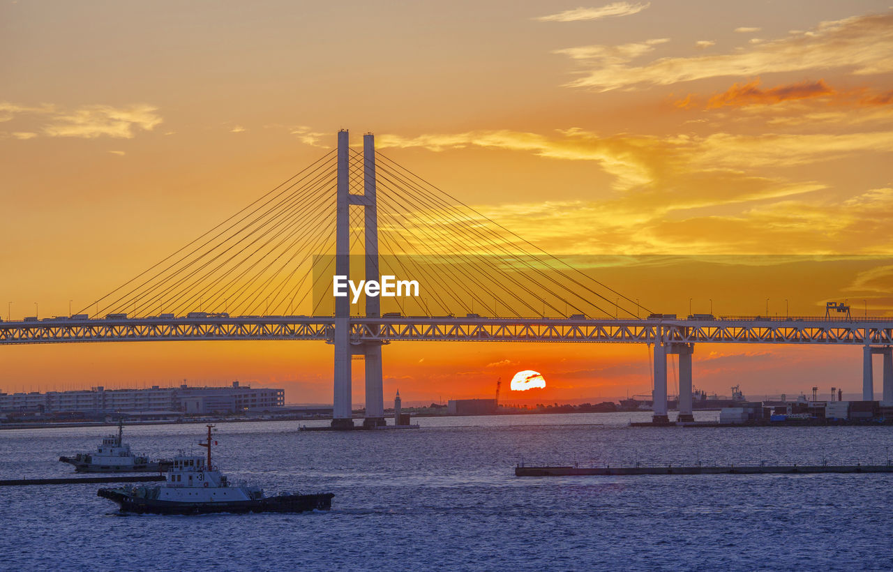 Boats in sea with cable-stayed bridge against orange sky during sunset