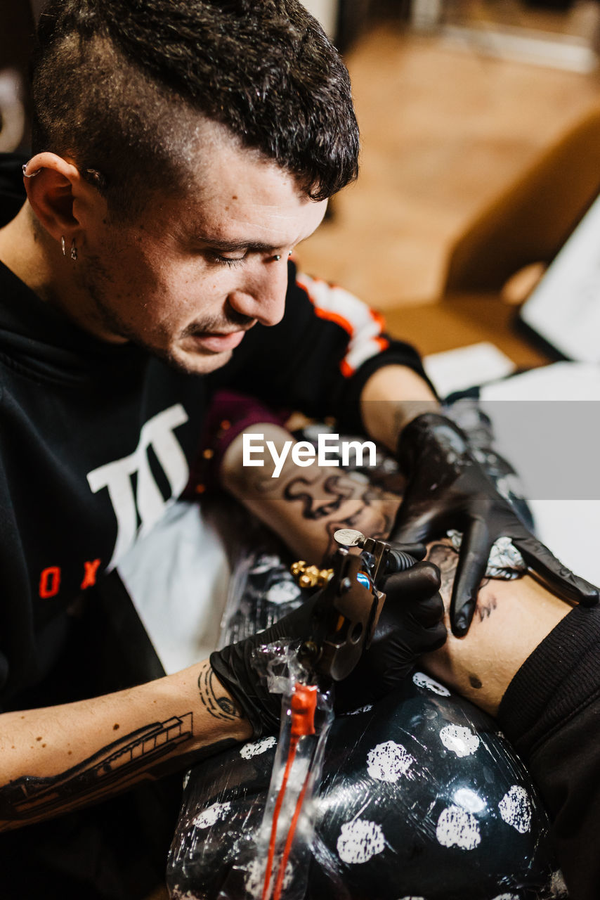 From above stylish man with piercing using tattoo machine to make tattoo on leg of crop customer during work in salon