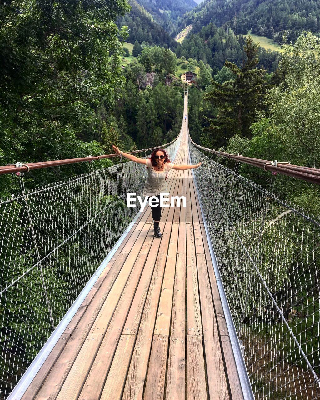 Woman with arms outstretched walking on footbridge against trees