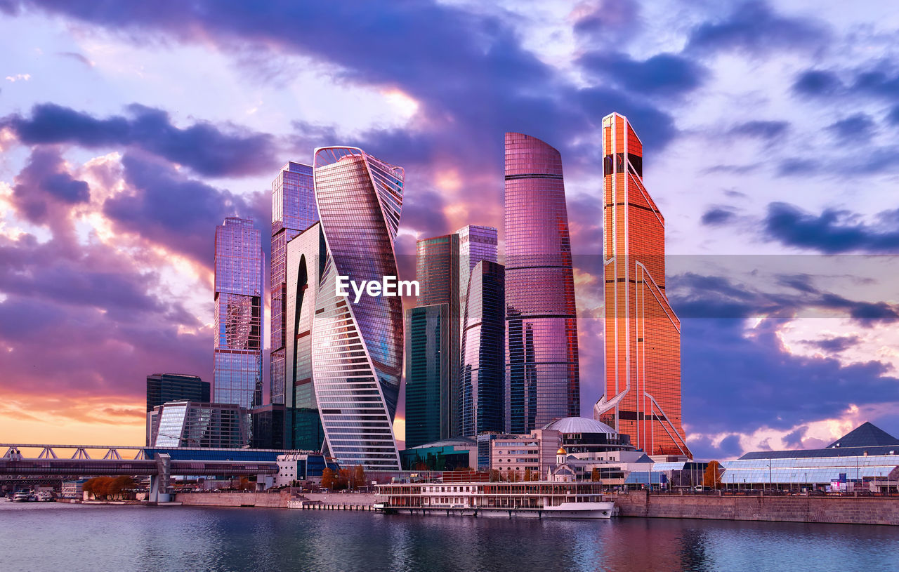 Modern buildings by river against sky during sunset