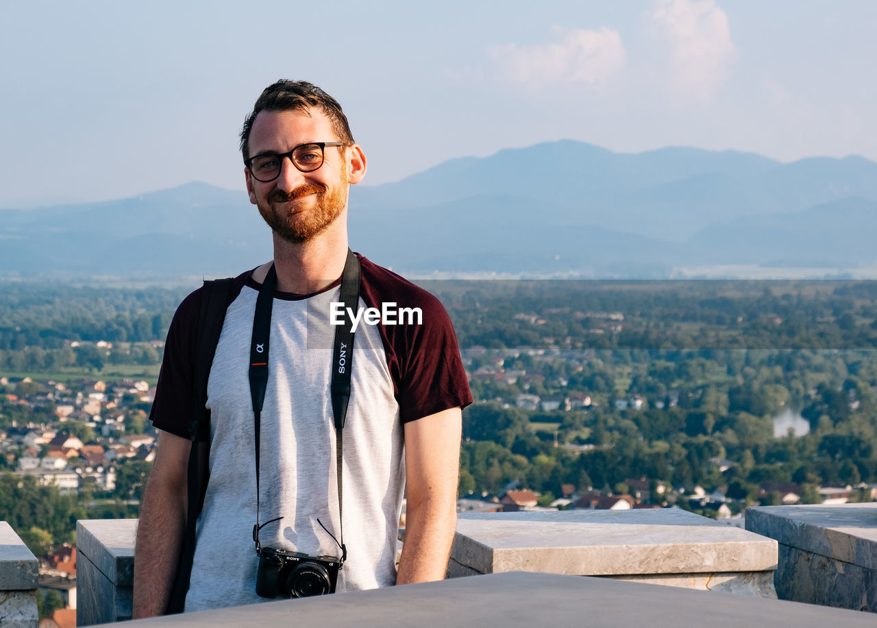 one person, adult, portrait, architecture, glasses, mountain, men, sky, beard, city, nature, facial hair, casual clothing, smiling, looking at camera, front view, vacation, standing, waist up, young adult, travel, building exterior, happiness, lifestyles, day, cityscape, built structure, leisure activity, emotion, fashion, travel destinations, landscape, person, outdoors, sunlight, eyeglasses, relaxation, sunglasses, spring, mountain range, clothing, copy space, building, holiday, t-shirt