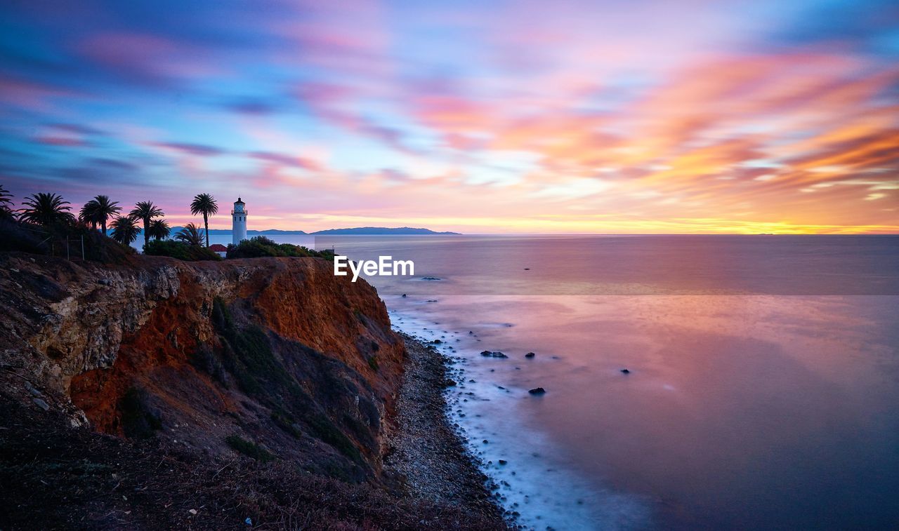 Scenic view of sea by cliff against cloudy sky during sunset