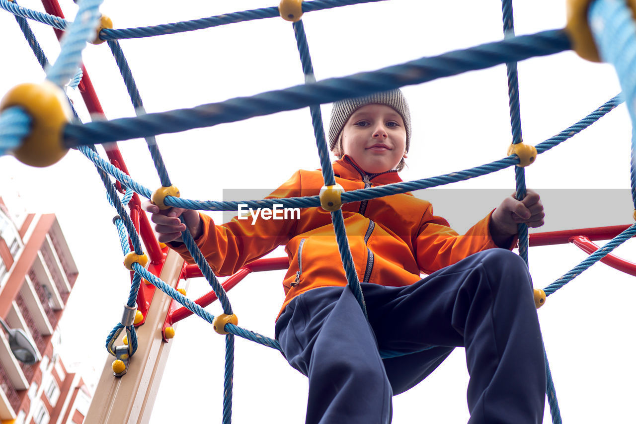Funny caucasian blond boy in an orange jacket plays on the sports playground