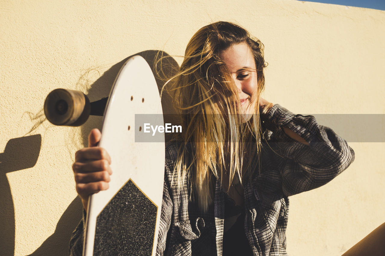 Smiling young woman with skateboard sitting against retaining wall on sunny day