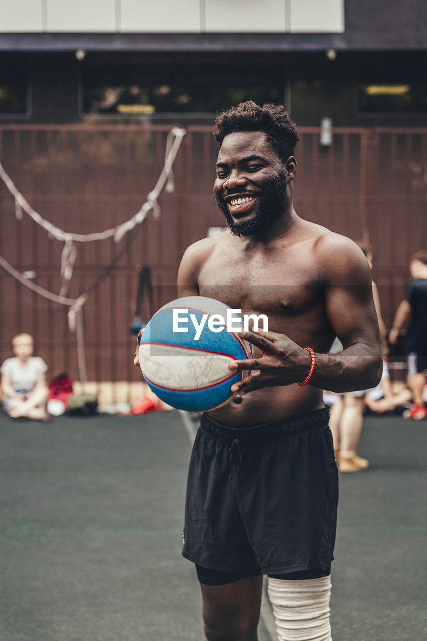 Happy shirtless man with sports ball standing at basketball court