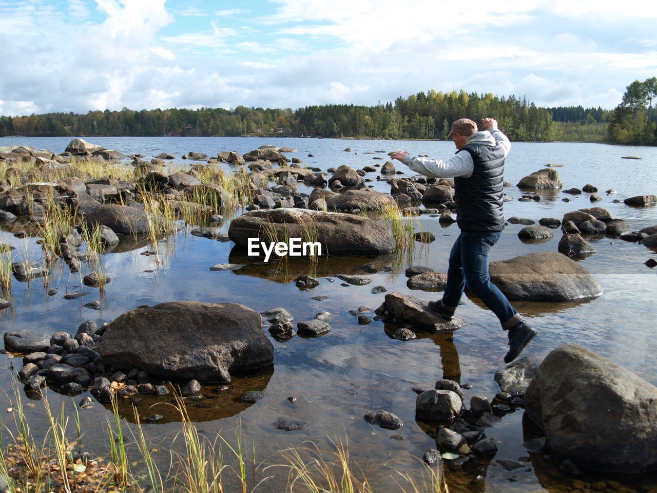 Rear view of playful man jumping on rocks in lake against cloudy sky