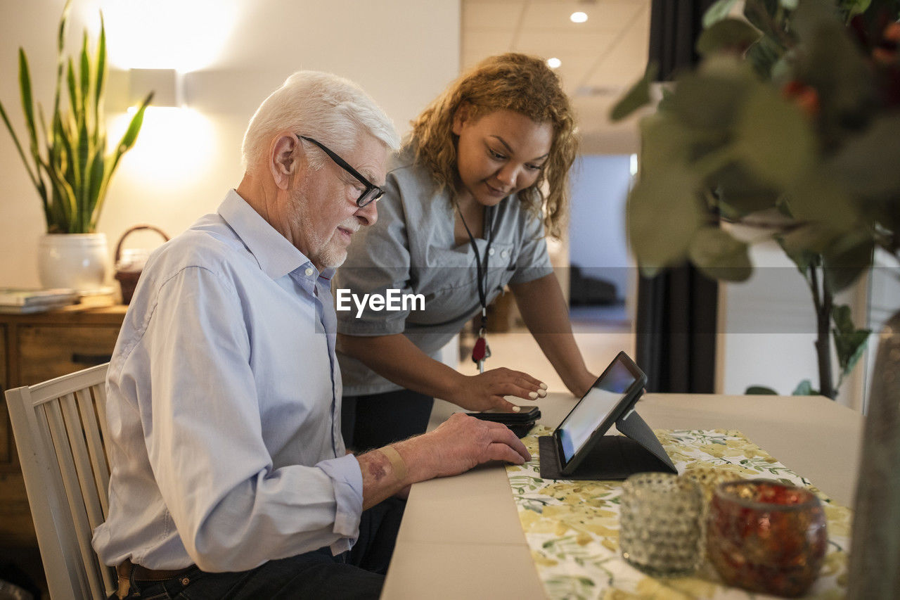 Young female caregiver assisting senior man in using digital tablet at retirement home
