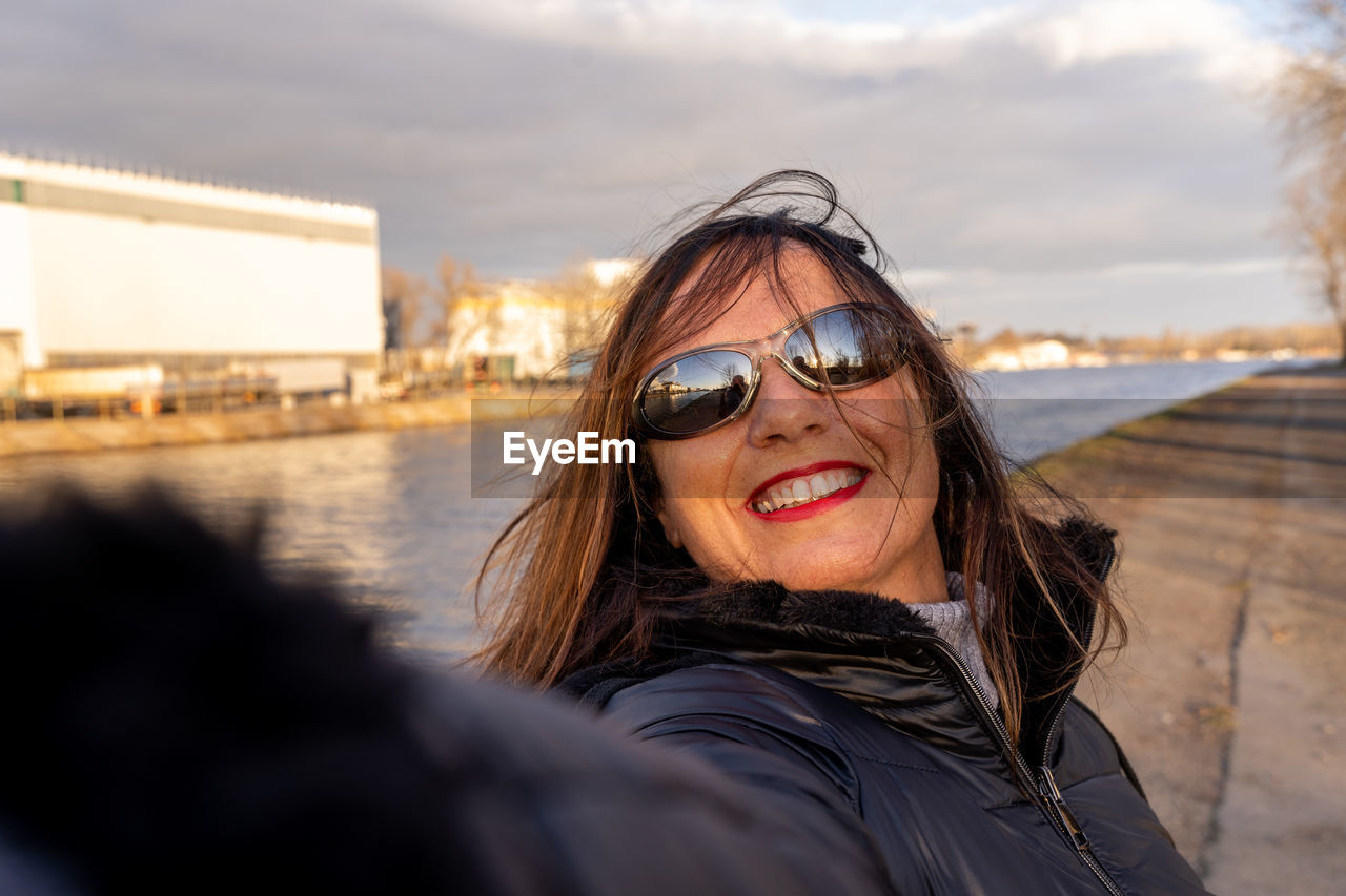 Middle aged woman wearing winter clothes taking selfie by a river - concept of people in recreation