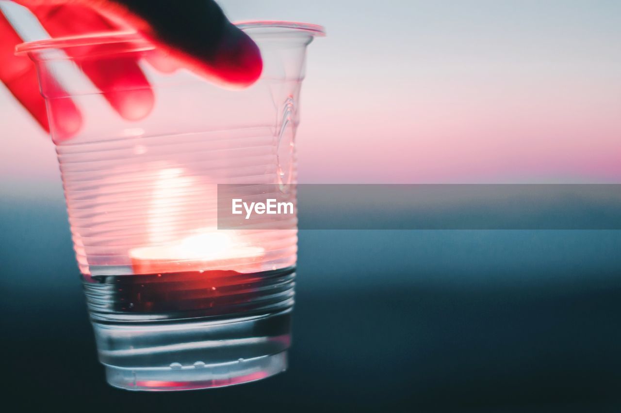 Cropped hand holding drink in disposable glass during sunset