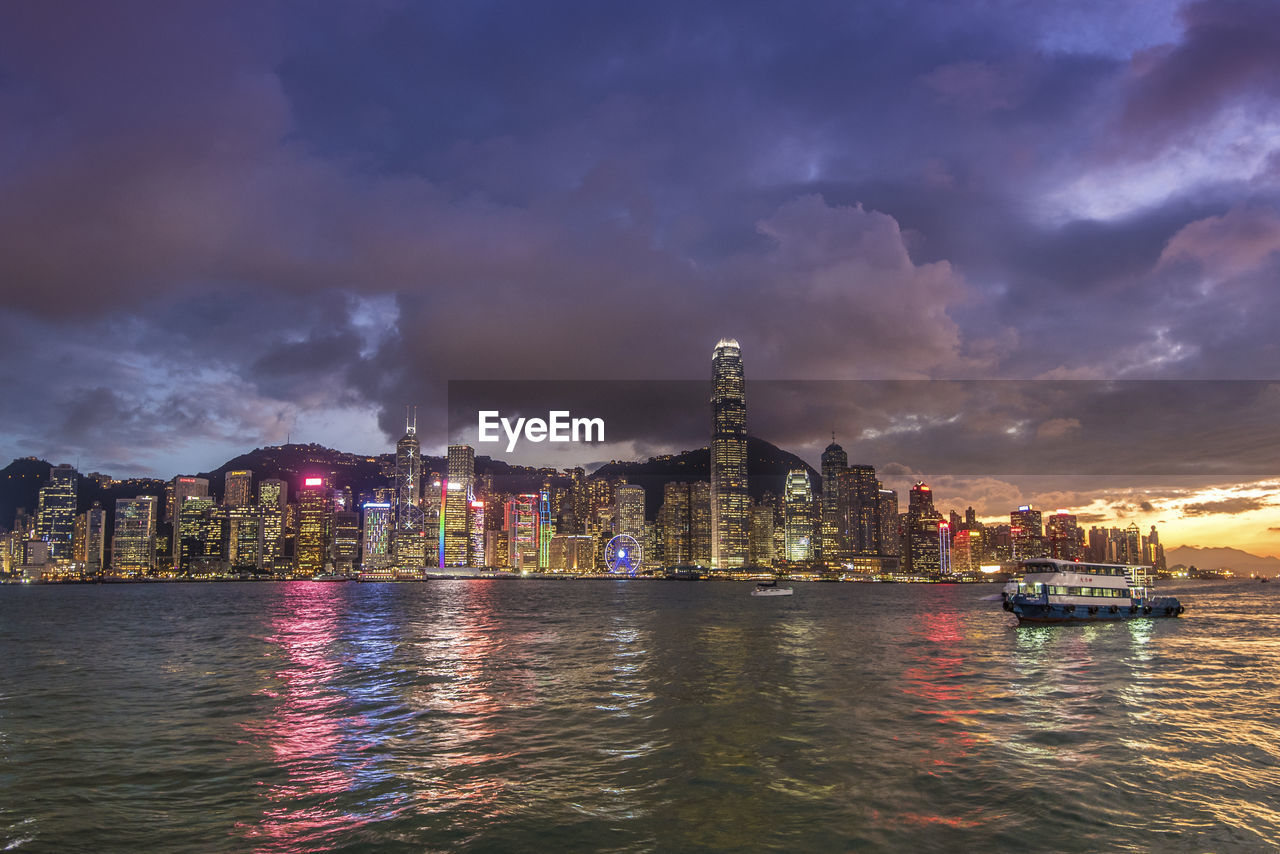 View of illuminated buildings by river against cloudy sky