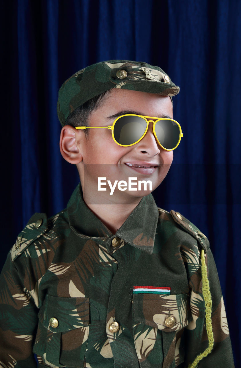 Smiling boy in army uniform and sunglasses standing against curtain