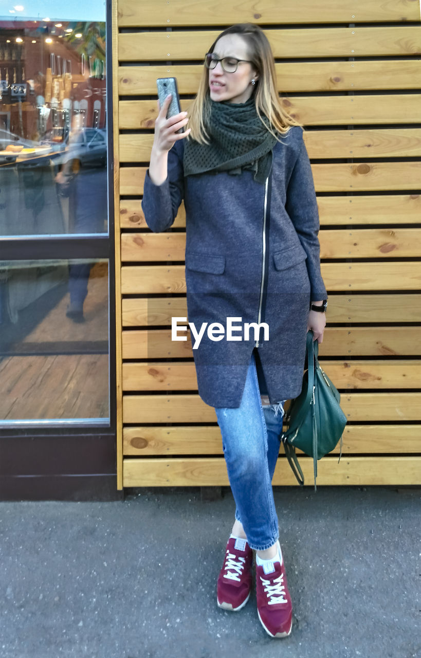 Young woman in coat and jeans indignantly looks at smartphone screen in the street near wooden wall
