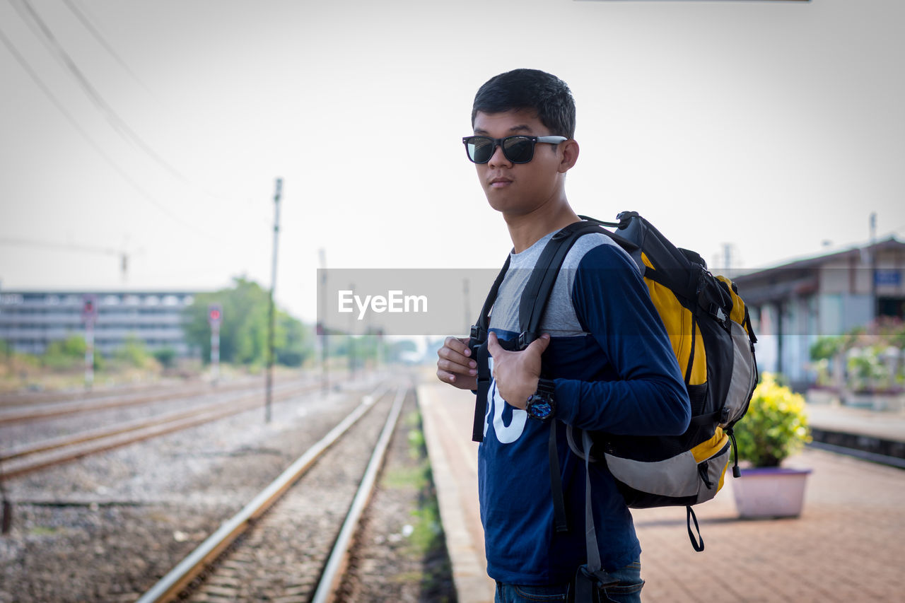 Young man wearing sunglasses while standing on railroad track against clear sky
