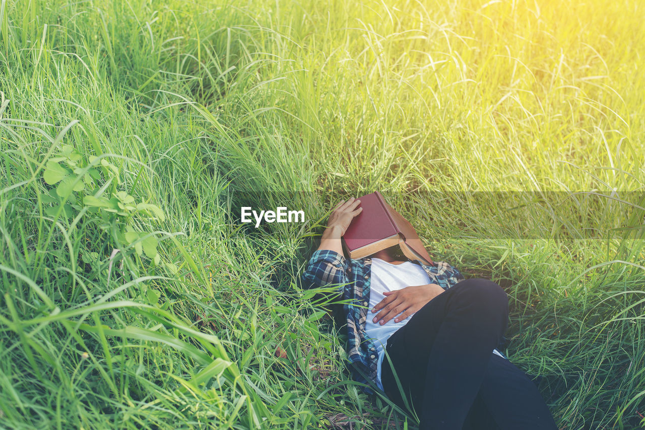 Man covering face with book while lying on grassy field