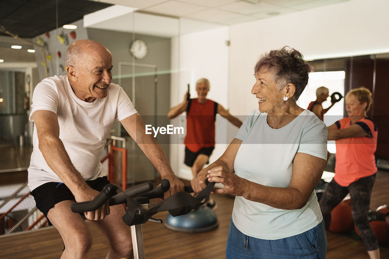 Cheerful senior woman in activewear laughing while standing near motivated husband exercising on cycling machine during workout in modern gym