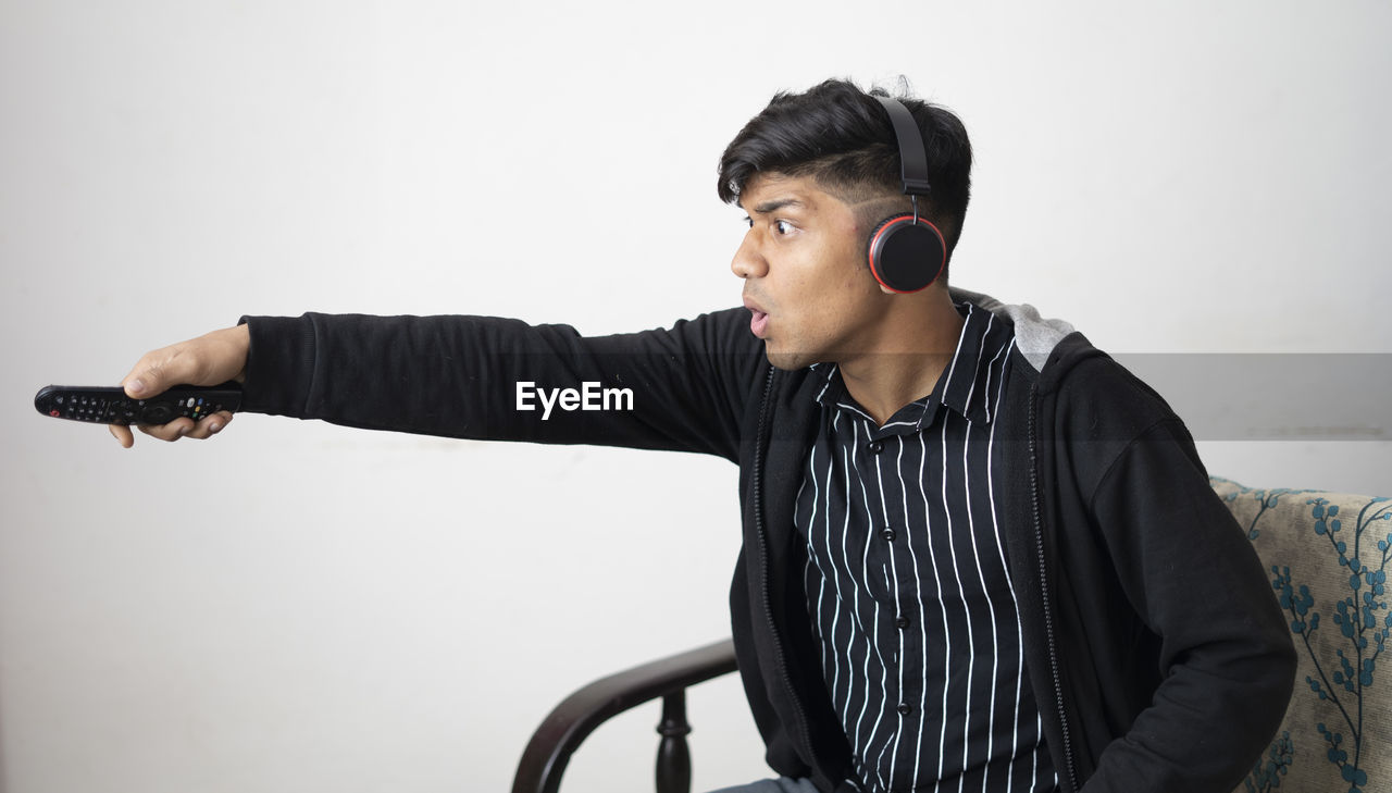 Asian man with headphone wearing black jacket holding remote excited to the match with arms raised.