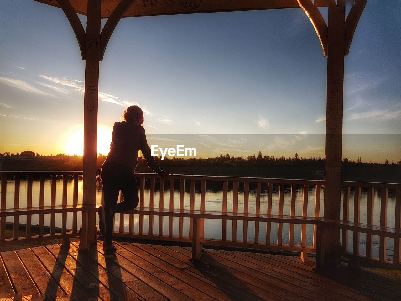Woman standing in gazebo by river against sky during sunset