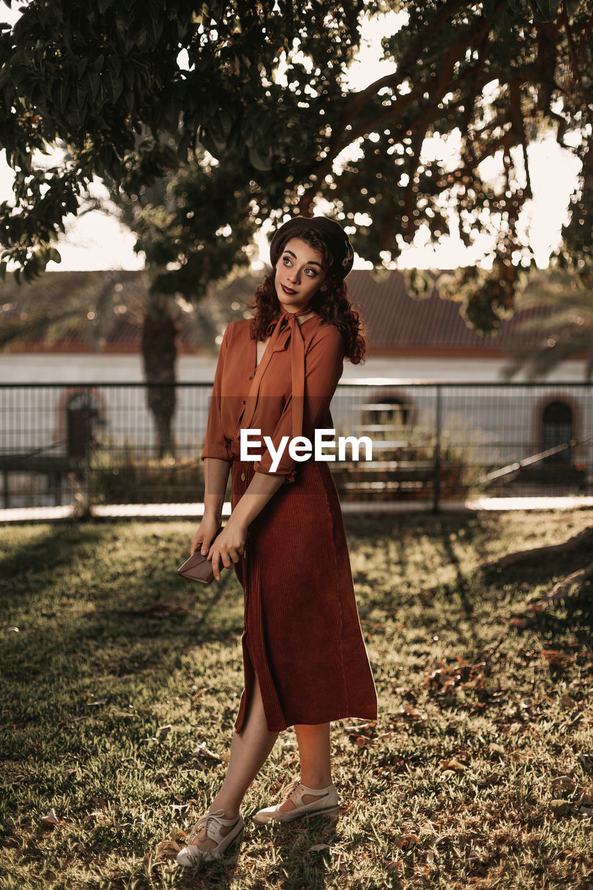 Brunette female in romantic vintage blouse and skirt standing in crossed graceful pose on grass holding book in hands while having pensive look