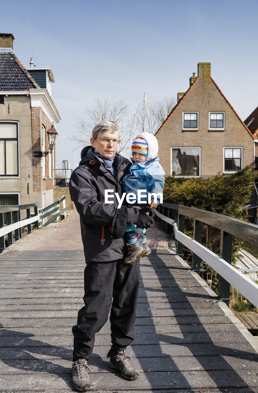 Portrait of grandfather carrying baby girl while standing on footbridge against houses