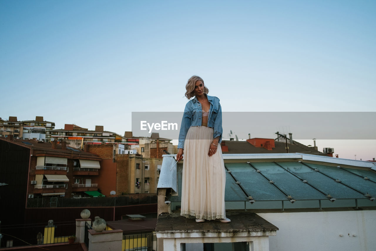 Tranquil female wearing chiffon skirt and denim jacket standing on roof of building and looking at camera against sundown sky