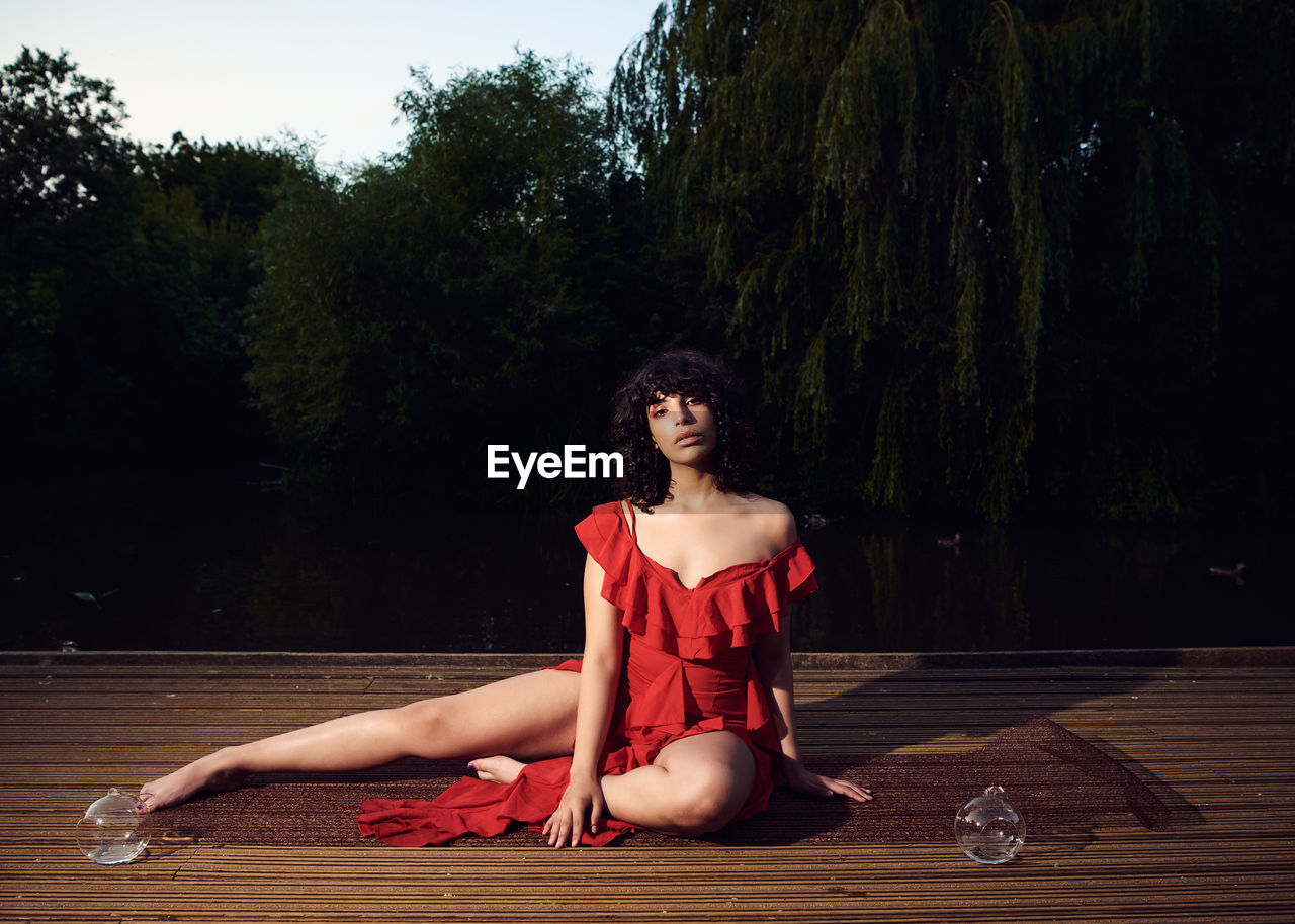 PORTRAIT OF YOUNG WOMAN SITTING ON WOOD AGAINST TREE