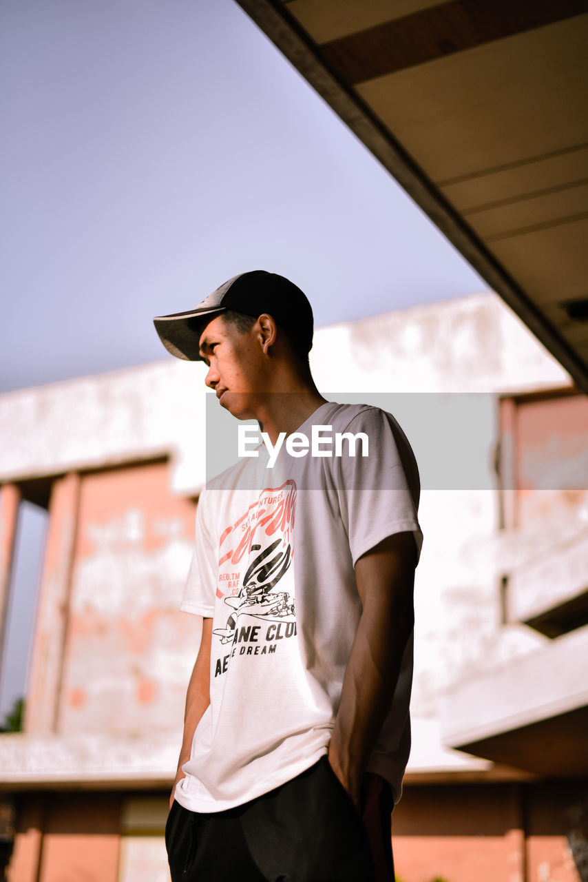 YOUNG MAN LOOKING AWAY WHILE STANDING AGAINST BUILDING