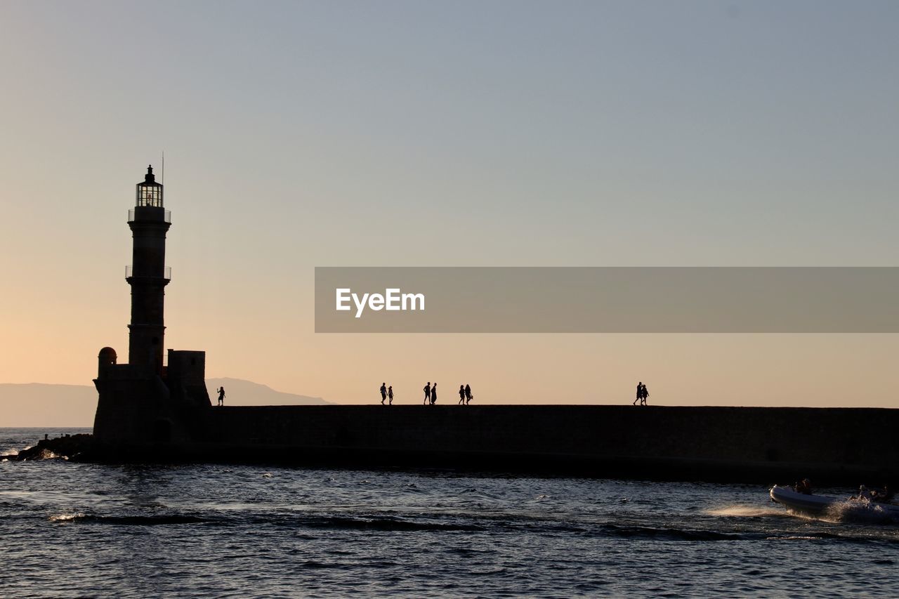 Silhouette people walking on pier towards lighthouse during sunset