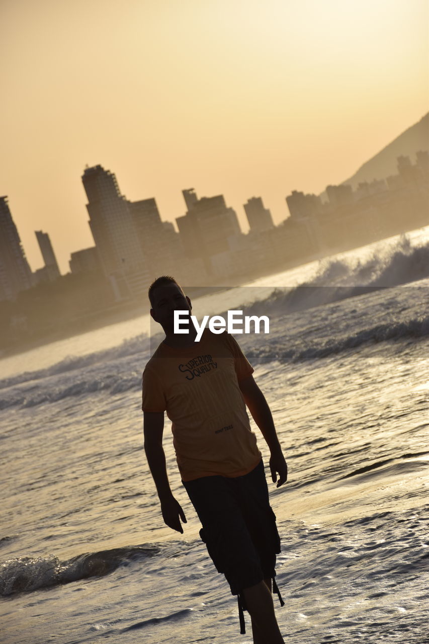 REAR VIEW OF MAN STANDING AT BEACH DURING SUNSET