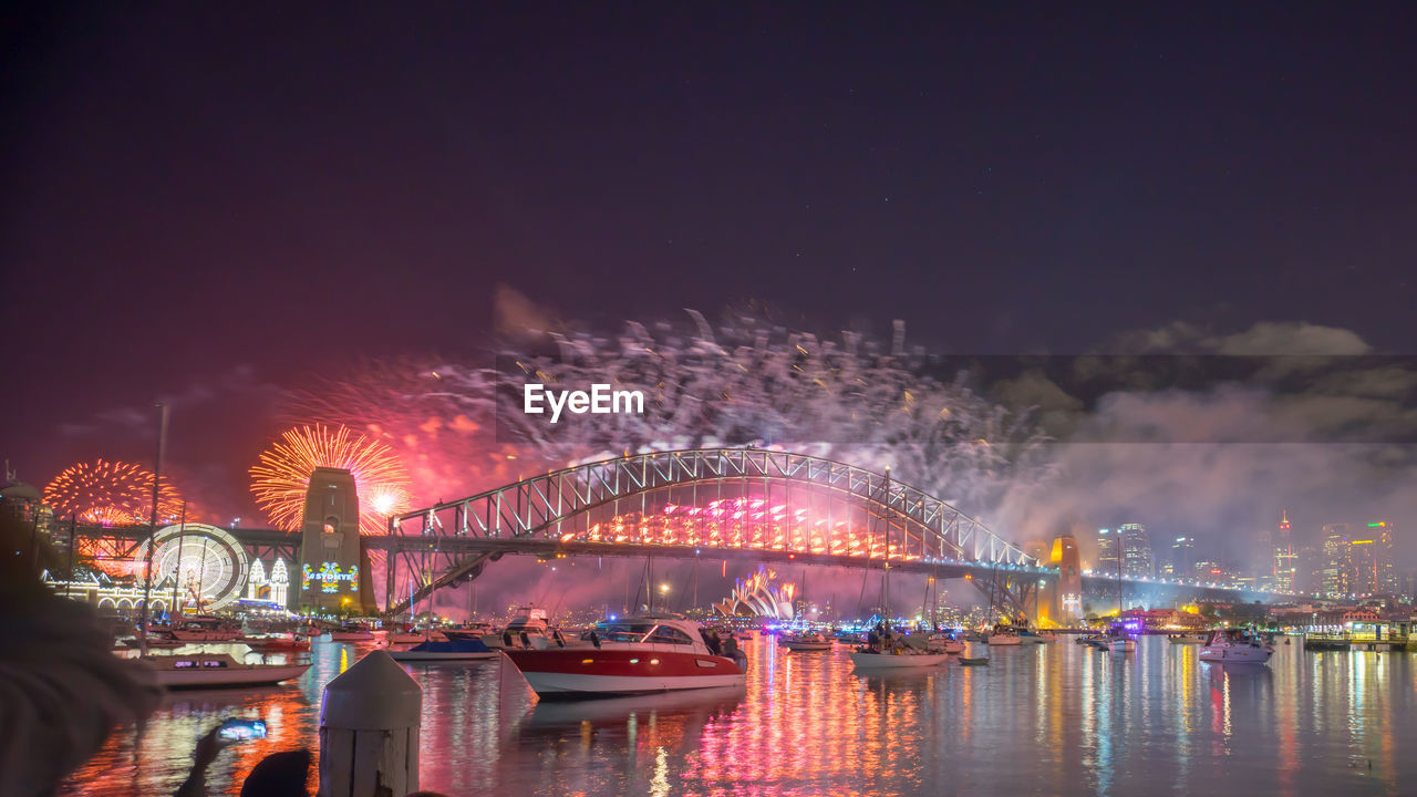 Low angle view of illuminated bridge over river with firework display in background at night